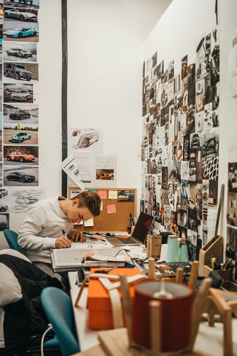 A designer sketching at a crowded studio desk under a wall of pinned car photos