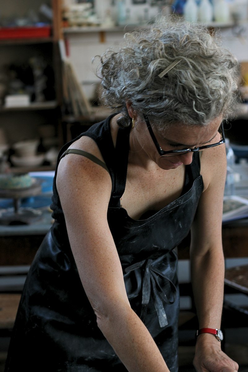 A potter in a work apron leaning over her bench in a busy studio